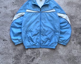 May include: A light blue zip-up track jacket featuring a white panel across the chest and shoulders with thin black piping. The jacket has a high collar, long sleeves with elastic cuffs, two zippered side pockets, and a small white Nike logo.