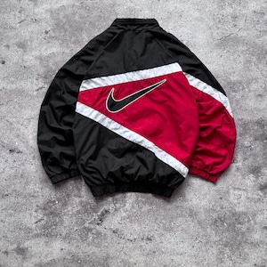 Op de afbeelding: Een zwart, rood en wit windjack met een groot zwart Nike swoosh-logo op de achterkant. Het jack heeft een hoge kraag, lange mouwen met elastische boorden en een color-block ontwerp met diagonale witte strepen die de secties van elkaar scheiden.
