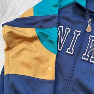 May include: A vintage track jacket in navy blue, teal, and gold. The jacket features a full zipper and the word "NIKE" in white block letters. The color-blocked design includes a teal collar and sleeve accents.