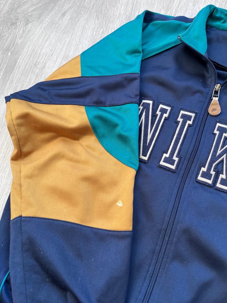 May include: A vintage track jacket in navy blue, teal, and gold. The jacket features a full zipper and the word "NIKE" in white block letters. The color-blocked design includes a teal collar and sleeve accents.