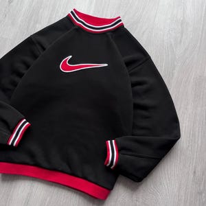 May include: Black sweatshirt with a red and white striped collar and cuffs. The sweatshirt features a red Nike swoosh logo outlined in white on the front. The bottom hem is red.