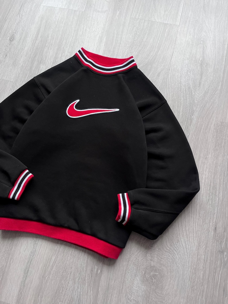 May include: Black sweatshirt with a red and white striped collar and cuffs. The sweatshirt features a red Nike swoosh logo outlined in white on the front. The bottom hem is red.