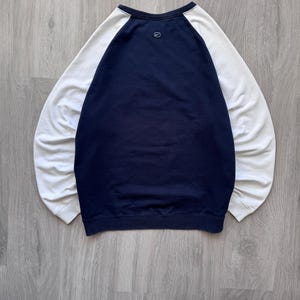May include: A navy blue and white raglan-sleeve sweatshirt. The body of the sweatshirt is navy blue, with white sleeves. A small Nike logo is visible on the upper back. The sweatshirt is laid flat on a wooden surface.