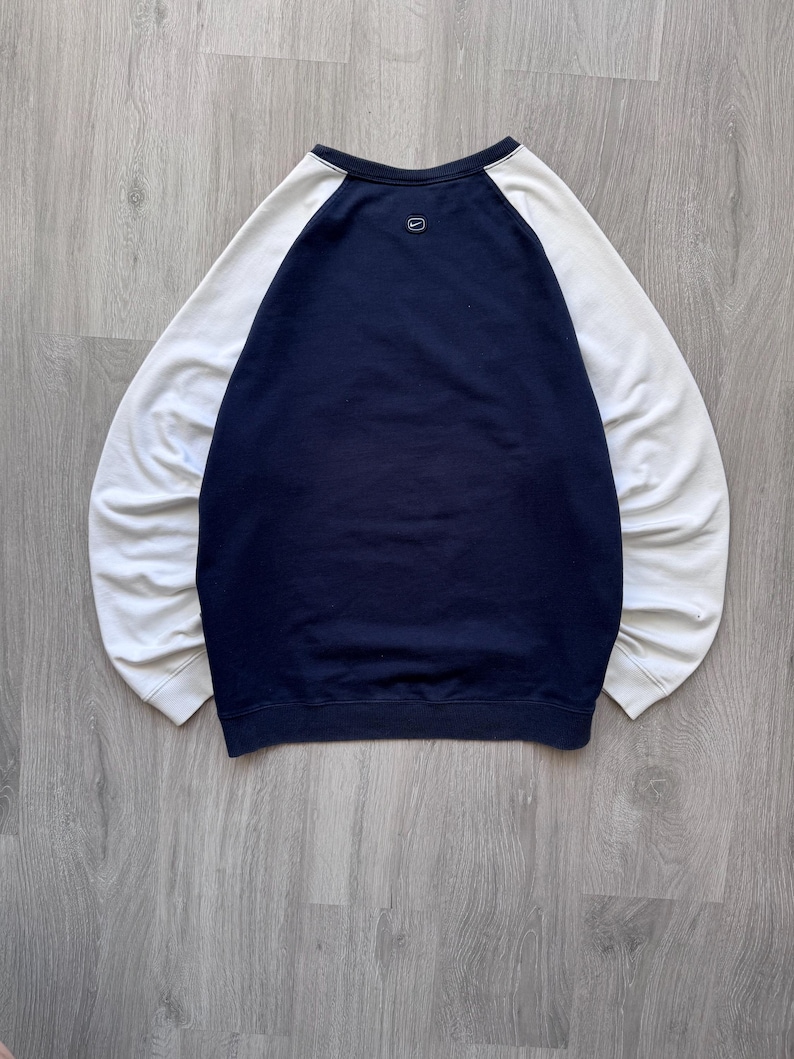May include: A navy blue and white raglan-sleeve sweatshirt. The body of the sweatshirt is navy blue, with white sleeves. A small Nike logo is visible on the upper back. The sweatshirt is laid flat on a wooden surface.
