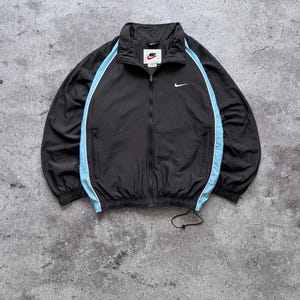 May include: A black zip-up track jacket featuring light blue piping along the sleeves and sides. The jacket has a high collar, a full-length front zipper, two zippered side pockets, and a white Nike swoosh logo on the left chest area.