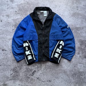 May include: A blue and black zip-up windbreaker jacket featuring a high collar and long sleeves. The front panels display the word NIKE in large white vertical lettering on black fabric strips. A small Nike logo is visible on the left sleeve.