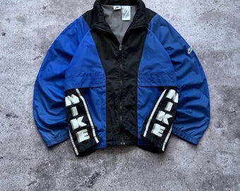 May include: A blue and black zip-up windbreaker jacket featuring a high collar and long sleeves. The front panels display the word NIKE in large white vertical lettering on black fabric strips. A small Nike logo is visible on the left sleeve.