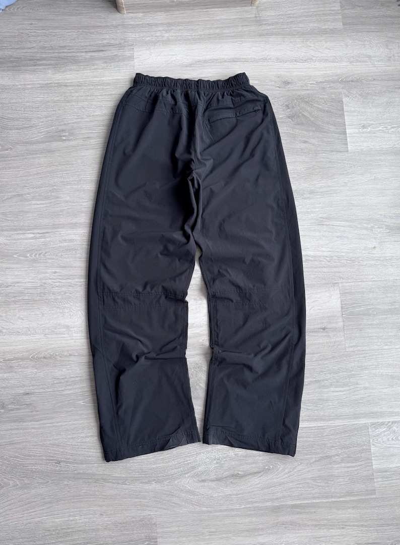 Puede incluir: Pantalones deportivos negros con cintura el&aacute;stica y un bolsillo trasero. Los pantalones est&aacute;n hechos de un material ligero y ligeramente brillante. Las perneras son rectas y anchas.