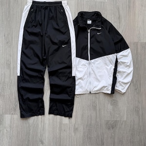 Peut inclure: Ensemble de surv&ecirc;tement noir et blanc. La veste est noire et blanche avec une fermeture &eacute;clair compl&egrave;te. Le pantalon est noir avec des rayures blanches sur les c&ocirc;t&eacute;s. Les deux pi&egrave;ces ont le logo Nike.