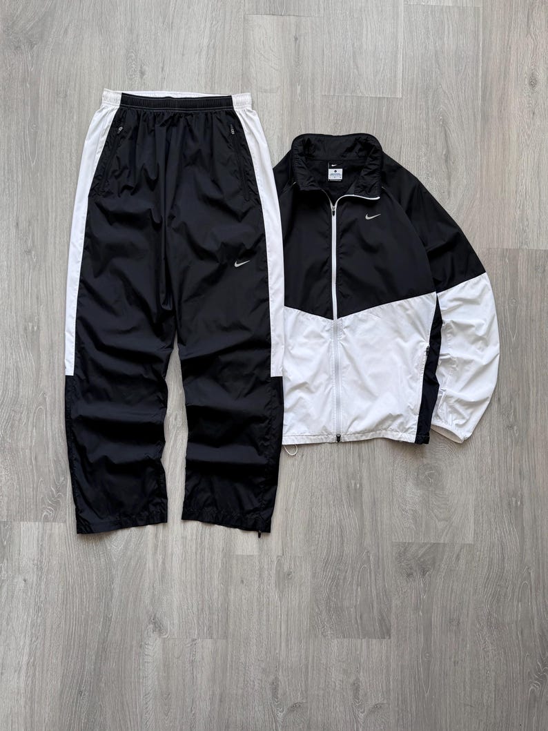 Peut inclure: Ensemble de surv&ecirc;tement noir et blanc. La veste est noire et blanche avec une fermeture &eacute;clair compl&egrave;te. Le pantalon est noir avec des rayures blanches sur les c&ocirc;t&eacute;s. Les deux pi&egrave;ces ont le logo Nike.
