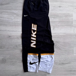以下が含まれることがあります： 左脚に白とゴールドの「NIKE」ロゴが入った黒色のナイキ製アスレチックパンツ。パンツの左下には白いナイキのスウッシュロゴがあり、白とゴールドのアクセントが施されています。