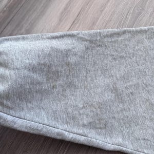 May include: Close-up of a gray fabric, possibly a section of a garment. The fabric has a textured appearance with visible stitching and a slight sheen. The color is a muted gray, and the fabric appears to be soft and flexible.