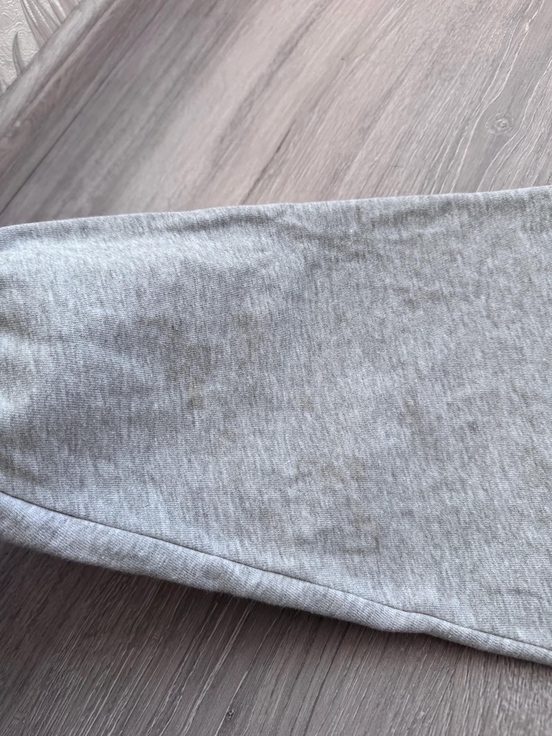 May include: Close-up of a gray fabric, possibly a section of a garment. The fabric has a textured appearance with visible stitching and a slight sheen. The color is a muted gray, and the fabric appears to be soft and flexible.