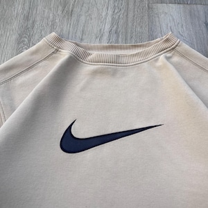 May include: A beige crew neck sweatshirt with a navy blue Nike swoosh logo embroidered on the chest. The sweatshirt has a ribbed neckline and a relaxed fit, suitable for casual wear.