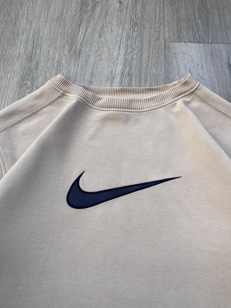 May include: A beige crew neck sweatshirt with a navy blue Nike swoosh logo embroidered on the chest. The sweatshirt has a ribbed neckline and a relaxed fit, suitable for casual wear.