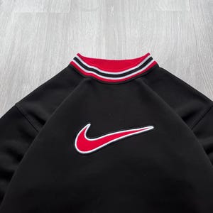 May include: Black sweatshirt featuring a red and white striped collar and a red Nike logo. The Nike logo has a white outline. The garment is made of a soft material.