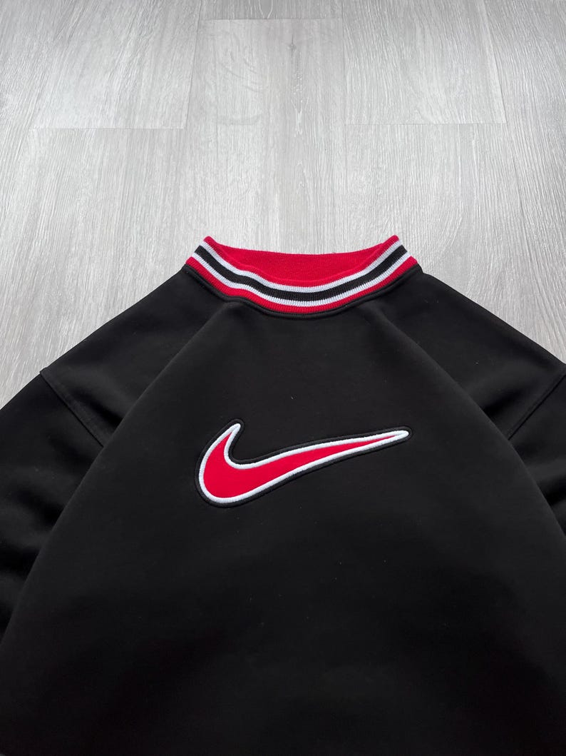 May include: Black sweatshirt featuring a red and white striped collar and a red Nike logo. The Nike logo has a white outline. The garment is made of a soft material.