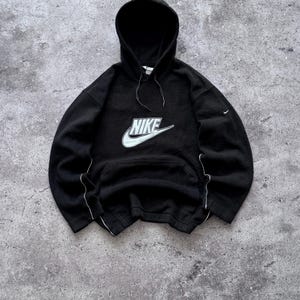 以下が含まれることがあります： 胸の中央にグレーの縁取りが施された大きな白いNikeロゴがあしらわれた、長袖の黒いフード付きスウェットシャツ。このパーカーには、フロントのカンガルーポケット、黒いドローストリング、袖に沿った細い白いパイピングのディテールが施されています。