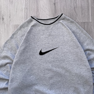 May include: A light gray sweatshirt with a black Nike swoosh logo on the chest. The crew neck has a black and white striped trim. The sweatshirt is made of a soft material.