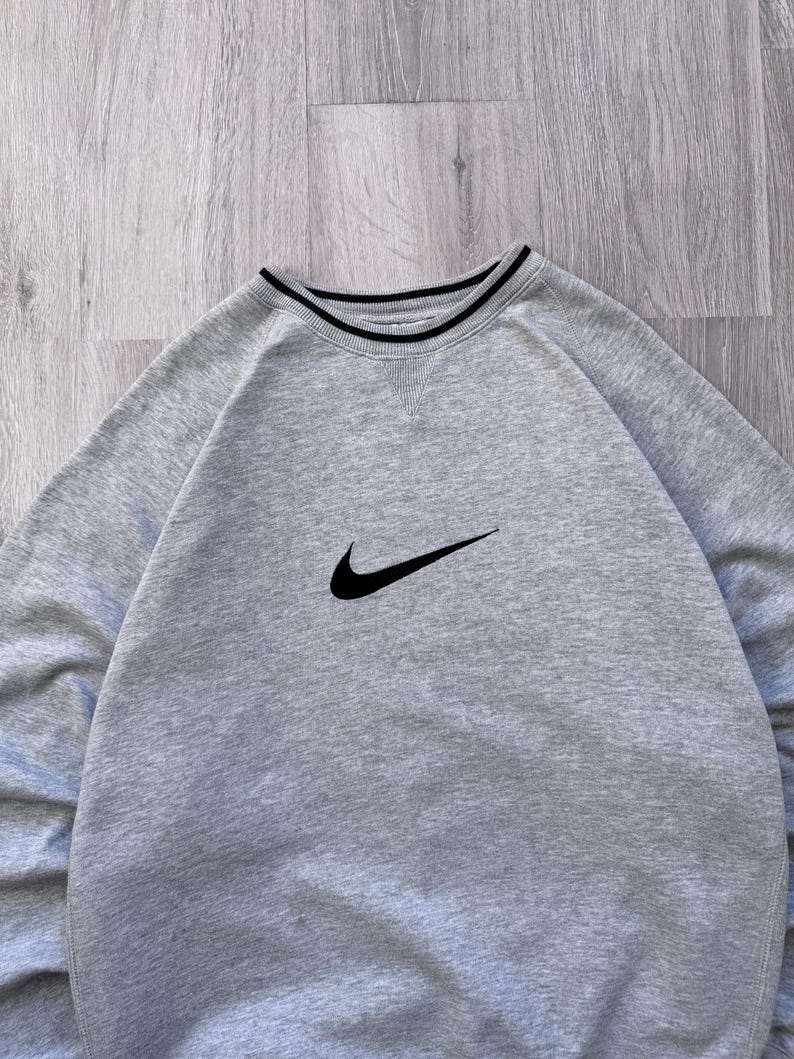 May include: A light gray sweatshirt with a black Nike swoosh logo on the chest. The crew neck has a black and white striped trim. The sweatshirt is made of a soft material.