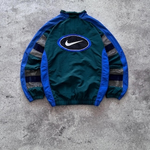 Peut inclure: Un coupe-vent vert foncé et bleu avec un col montant, des manches longues à empiècements en maille et un grand écusson ovale noir au dos portant le logo Nike blanc. La veste présente des poignets et un ourlet élastiques.