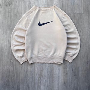 May include: A beige crew neck sweatshirt with a navy blue Nike swoosh logo on the chest. The sweatshirt has ribbed cuffs and hem. The garment is made of a soft material.