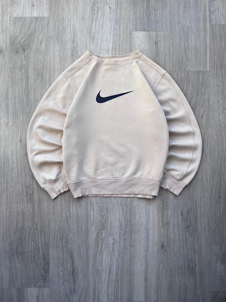 May include: A beige crew neck sweatshirt with a navy blue Nike swoosh logo on the chest. The sweatshirt has ribbed cuffs and hem. The garment is made of a soft material.
