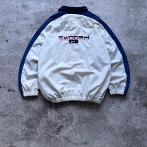 May include: A cream-colored windbreaker jacket featuring dark blue sleeves and a collar. The back displays the word SWOOSH in dark red lettering above a small dark blue rectangle containing a white Nike swoosh logo. The jacket has elastic cuffs.
