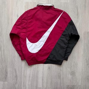 Könnte beinhalten: Rote, schwarze und weiße Nike Windjacke. Die Jacke hat ein großes weißes Nike Swoosh-Logo auf dem Rücken, mit einem roten Oberteil und einem schwarzen Unterteil. Der Kragen ist silberfarben.