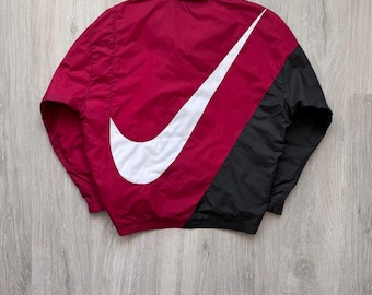 May include: A red, black, and white Nike windbreaker jacket. The jacket features a large white Nike swoosh logo on the back, with a red upper and black lower section. The collar is silver.