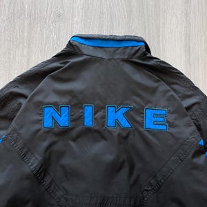 May include: Black Nike jacket with blue accents. The word "NIKE" is prominently displayed in blue block letters on the back. The jacket has a collar and a blue stripe along the top.