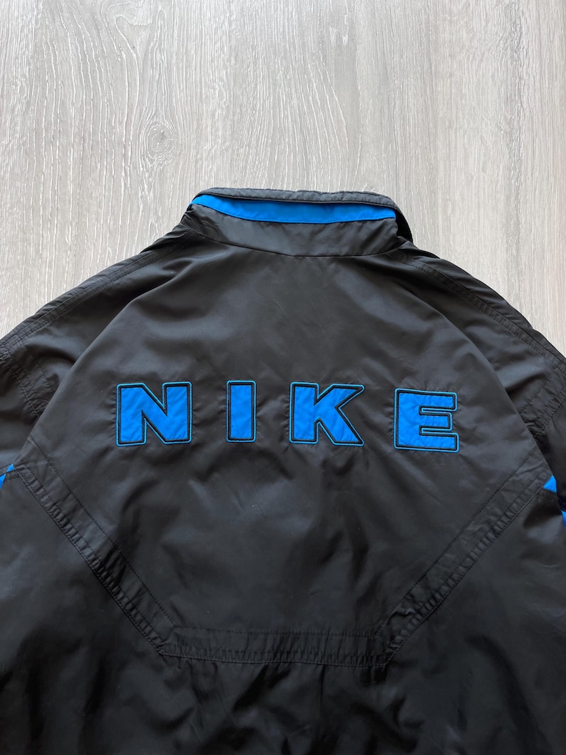 May include: Black Nike jacket with blue accents. The word "NIKE" is prominently displayed in blue block letters on the back. The jacket has a collar and a blue stripe along the top.