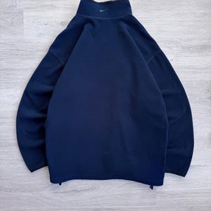 May include: Navy blue fleece pullover with a high collar and a small teal logo on the chest. The pullover has a black trim around the collar and cuffs. The material appears soft and warm, suitable for cooler weather.