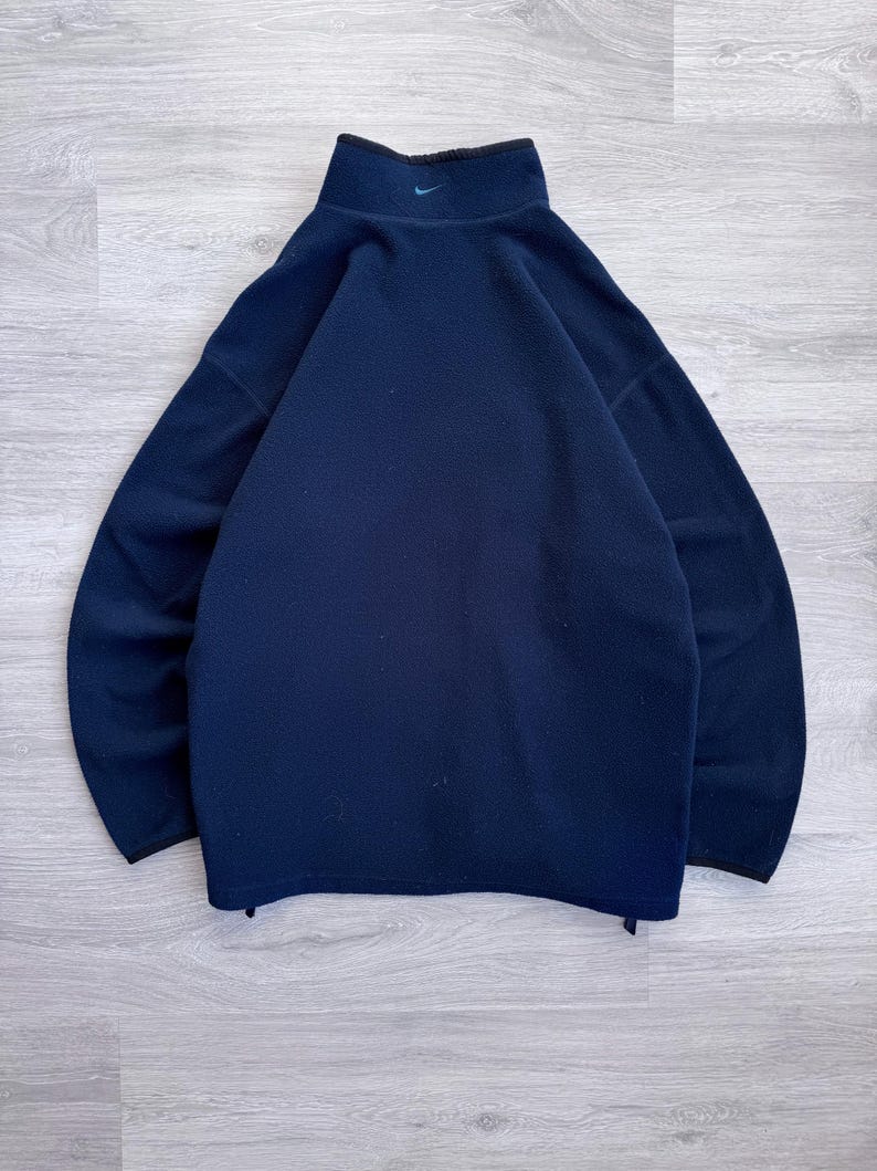 May include: Navy blue fleece pullover with a high collar and a small teal logo on the chest. The pullover has a black trim around the collar and cuffs. The material appears soft and warm, suitable for cooler weather.