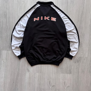 May include: A black track jacket featuring white panels along the sleeves and a high collar. The back displays the word NIKE in bold, orange-outlined white lettering. The garment has a ribbed hem and cuffs, showcasing a classic athletic design.