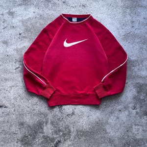 Può includere: Una felpa rossa a maniche lunghe con il logo Nike swoosh bianco sul petto. Il capo presenta un girocollo a coste con una sottile riga bianca, polsini a coste e un orlo a coste. Un sottile profilo bianco corre lungo la parte esterna delle maniche.