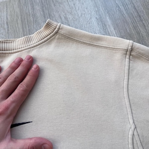 May include: A beige crew neck sweatshirt with a small black logo on the left chest. The sweatshirt has a ribbed collar and cuffs, and visible stitching details. The fabric appears to be a soft, knit material.