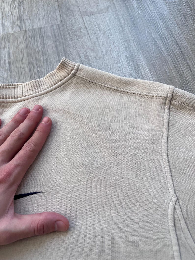May include: A beige crew neck sweatshirt with a small black logo on the left chest. The sweatshirt has a ribbed collar and cuffs, and visible stitching details. The fabric appears to be a soft, knit material.
