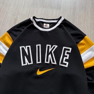Puede incluir: Sudadera negra Nike con letras "NIKE" en blanco con contorno y un logotipo de swoosh amarillo. Las mangas tienen rayas amarillas y blancas. El cuello y los pu&ntilde;os son blancos.