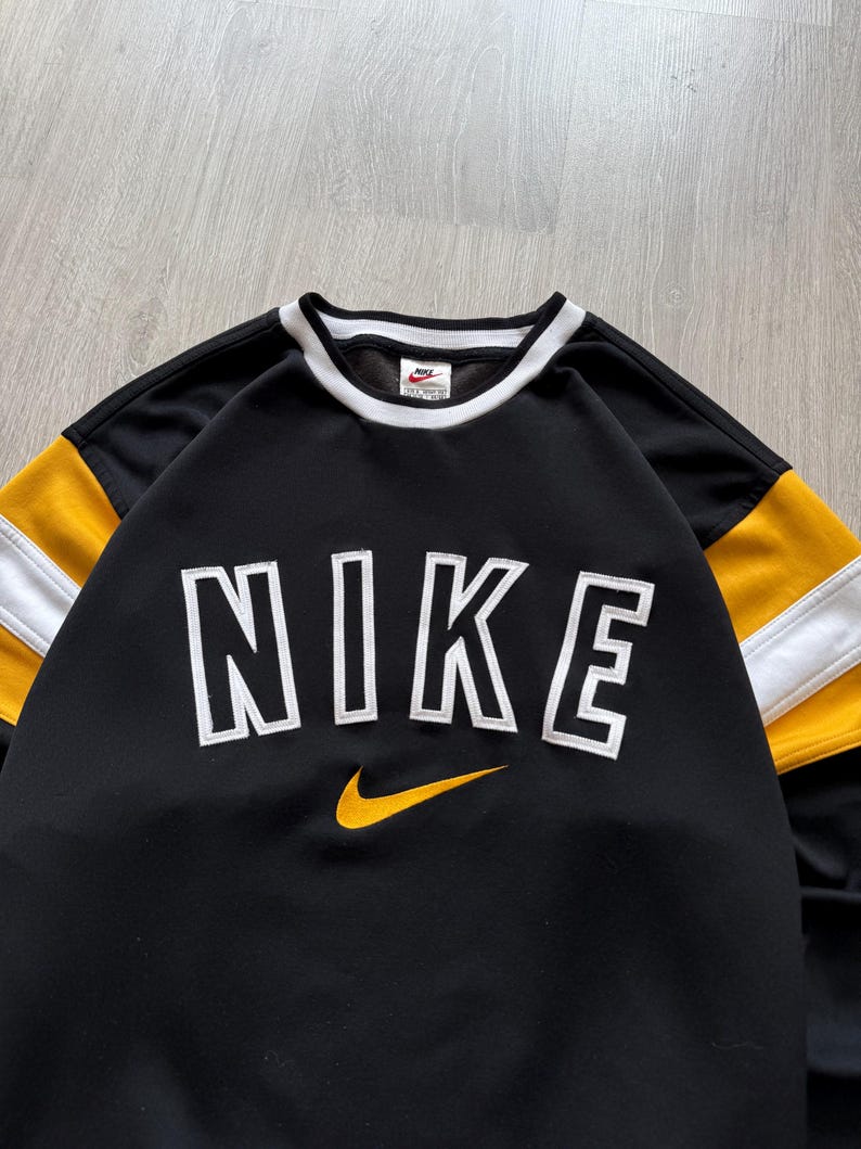 Puede incluir: Sudadera negra Nike con letras "NIKE" en blanco con contorno y un logotipo de swoosh amarillo. Las mangas tienen rayas amarillas y blancas. El cuello y los pu&ntilde;os son blancos.