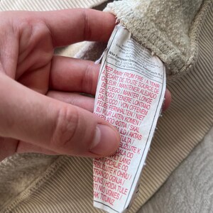 May include: Close-up of a beige sweatshirt's interior, showcasing the textured fabric and a sewn-in tag. The tag features multi-language safety warnings and the Nike European Operations B.V. logo. The sweatshirt's ribbed texture is visible.