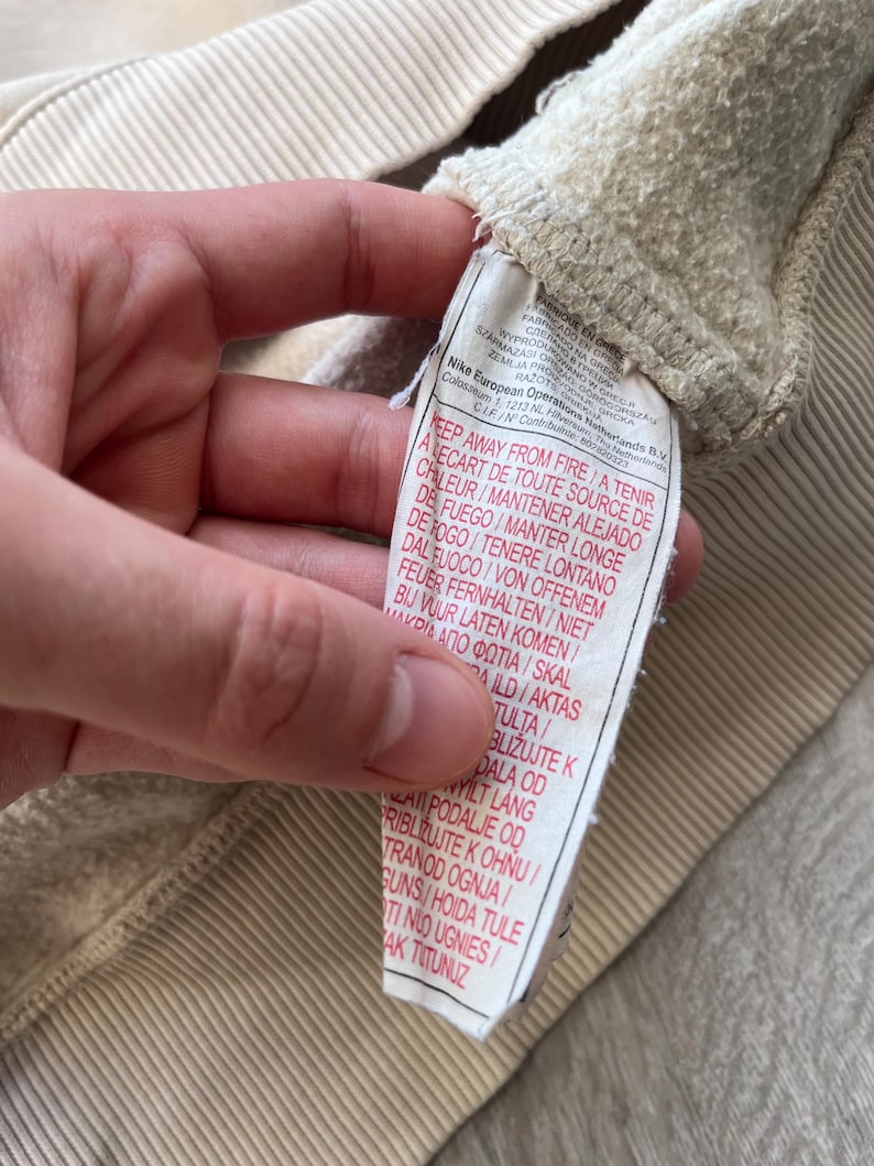 May include: Close-up of a beige sweatshirt's interior, showcasing the textured fabric and a sewn-in tag. The tag features multi-language safety warnings and the Nike European Operations B.V. logo. The sweatshirt's ribbed texture is visible.