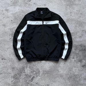 May include: A black zip-up track jacket featuring a white horizontal stripe across the chest and shoulders, with matching white vertical stripes running down the length of each sleeve. A small black Nike swoosh logo is embroidered on the chest.