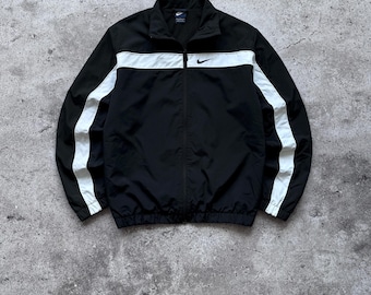May include: A black zip-up track jacket featuring a white horizontal stripe across the chest and shoulders, with matching white vertical stripes running down the length of each sleeve. A small black Nike swoosh logo is embroidered on the chest.