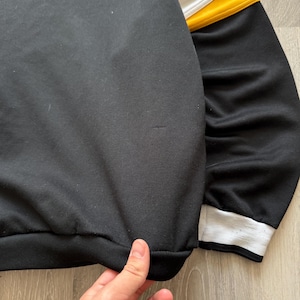 Puede incluir: Sudadera negra con rayas amarillas y blancas en la parte superior de la manga. El pu&ntilde;o es blanco con una raya negra. El dobladillo inferior es negro. La tela parece ser un material suave y c&oacute;modo.