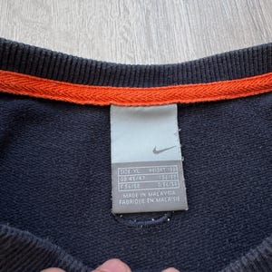 May include: Navy blue sweatshirt with an orange trim at the neckline. A tag with the Nike logo and size information is visible. The tag includes size XL, height 189, and other size details.