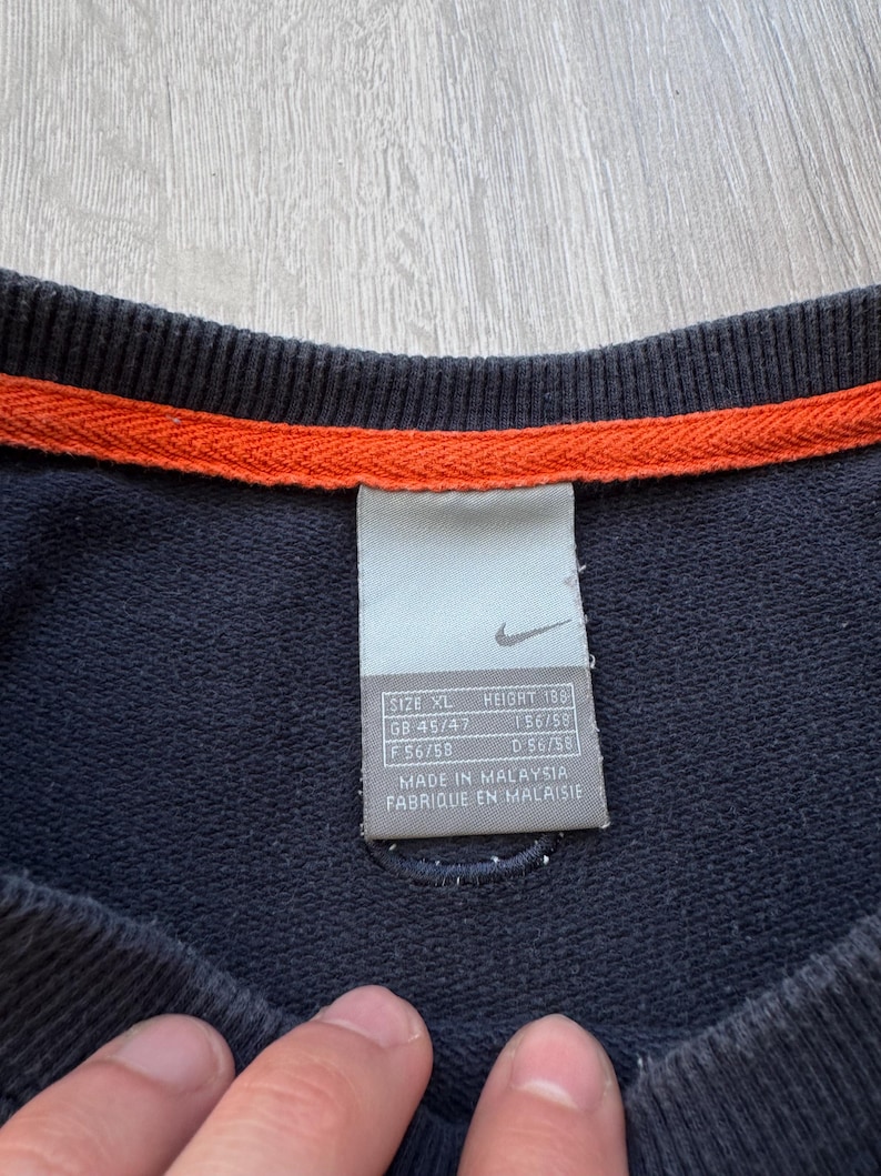 May include: Navy blue sweatshirt with an orange trim at the neckline. A tag with the Nike logo and size information is visible. The tag includes size XL, height 189, and other size details.