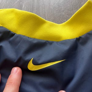 May include: Close-up of a navy blue jacket with a yellow collar and the Nike logo. The jacket has a yellow embroidered Nike swoosh on the front. The collar is a ribbed knit fabric.