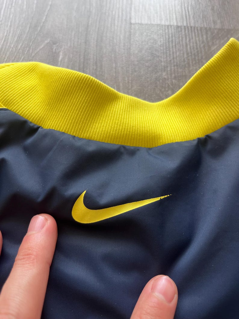 May include: Close-up of a navy blue jacket with a yellow collar and the Nike logo. The jacket has a yellow embroidered Nike swoosh on the front. The collar is a ribbed knit fabric.