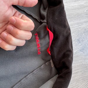 May include: Close-up of a gray and black fleece jacket. The jacket has a red pull tab and the word "TOM MAX" embroidered in red on the gray fabric. The jacket is made of a soft, textured material.